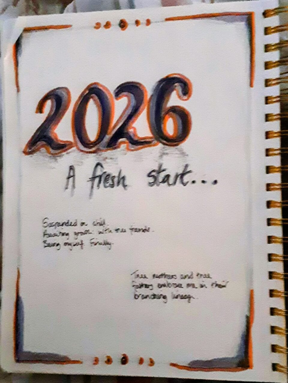 A spiral bound notebook page with the words '2026 a fresh start' drawn in black grey and orange pen.