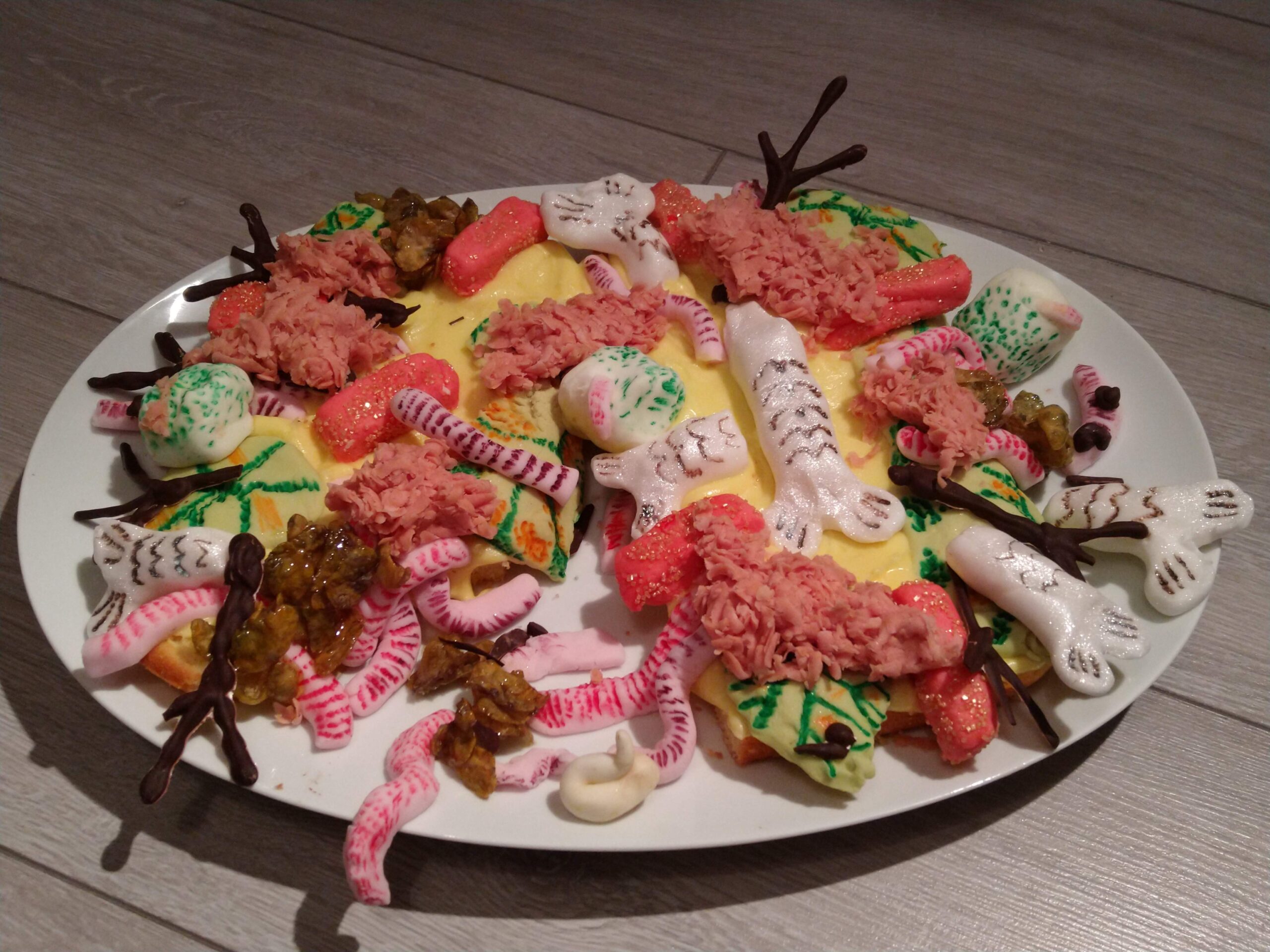 A homemade cake inspired by Mr Twit's revolting sandwich from Roald Dahl's The Twits, piled onto a white oval platter and decorated with pink icing worms, white sugar bones, chocolate bird feet, brightly coloured sweets and lumpy brown and pink toppings to resemble a disgusting mixture of ingredients.