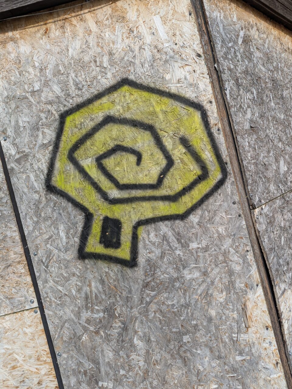Close-up of a yellow graffiti symbol formed from a black spiral on a boarded wall.