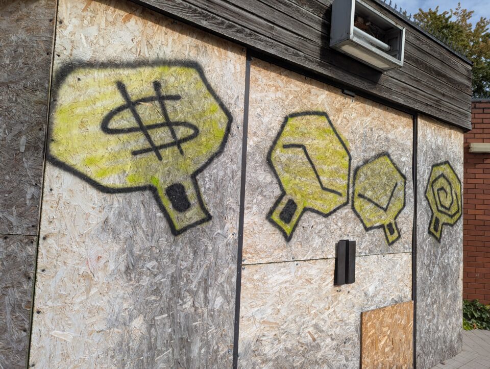 Oblique view of four yellow and black graffiti symbols across a boarded building.
