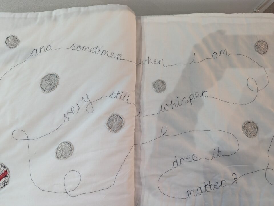 White textile artwork with looping stitched handwriting reading “and sometimes when I am very still I whisper does it matter?” and small circular fabric forms.