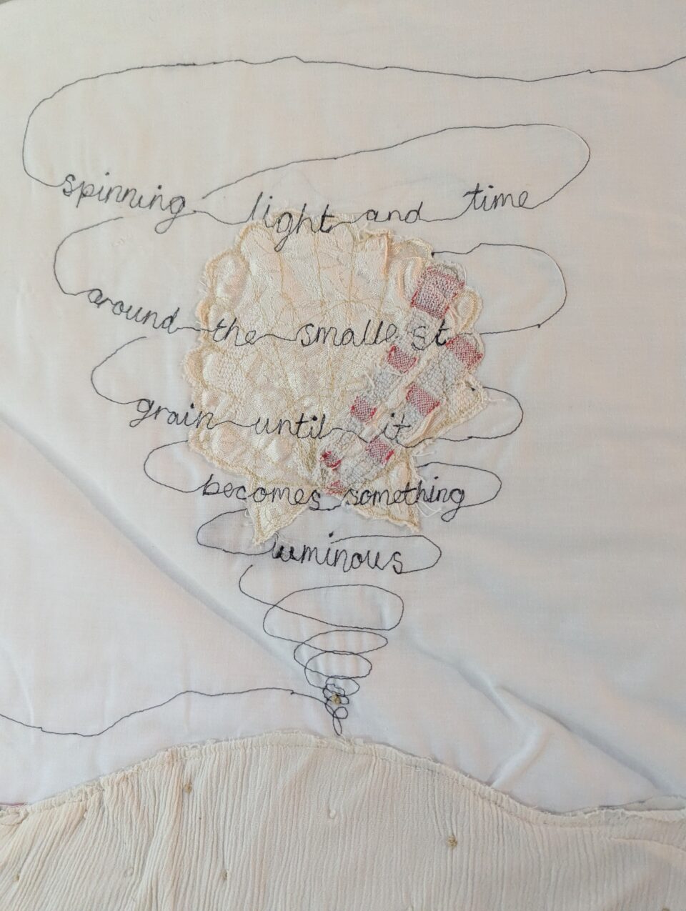 Layered textile artwork with a small patchwork figure and spiralling stitched text reading “spinning light and time around the smallest grain until it becomes something luminous”.