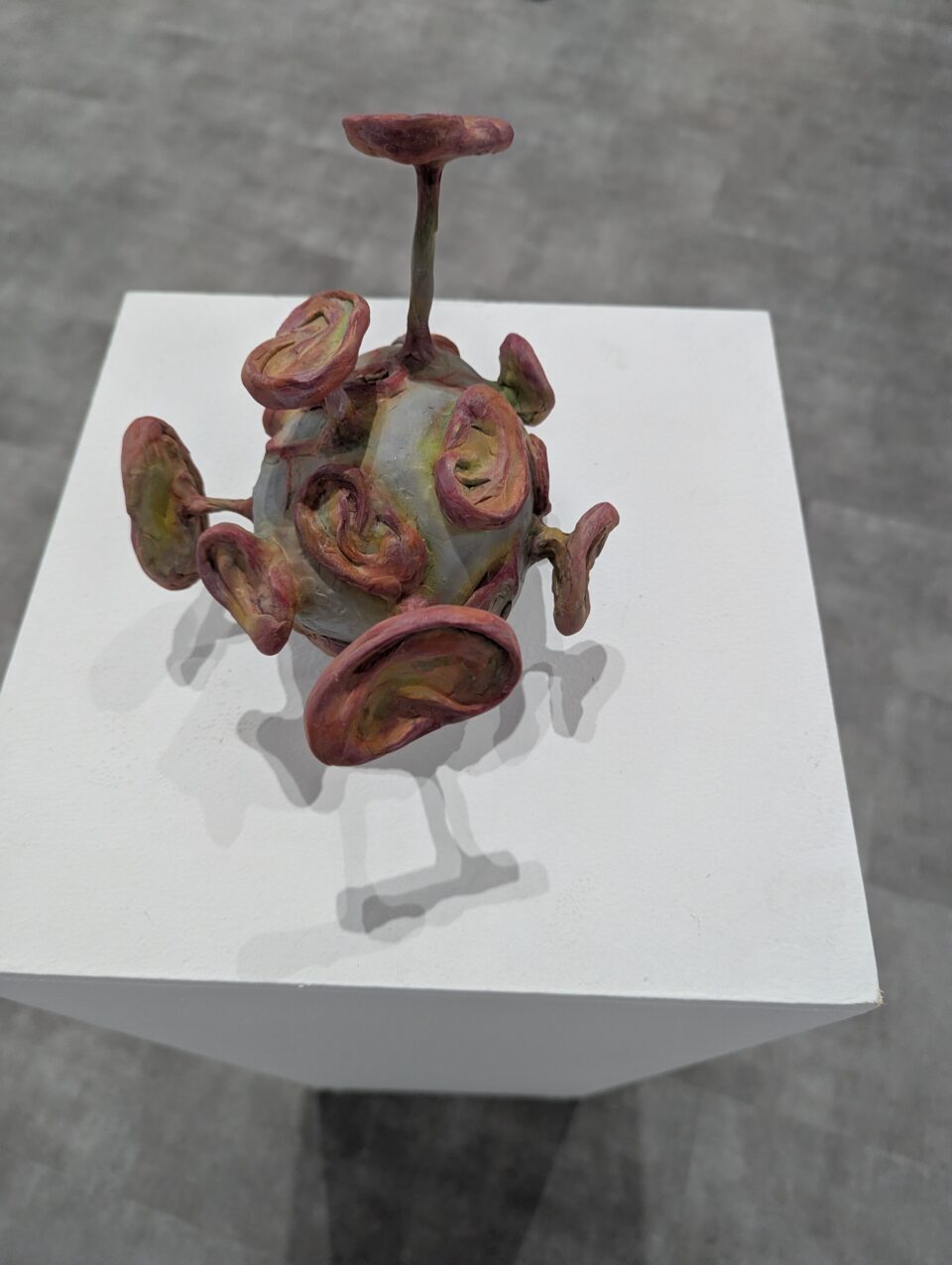Small sculptural form with a rounded body and multiple reddish disc-like projections displayed on a white plinth.