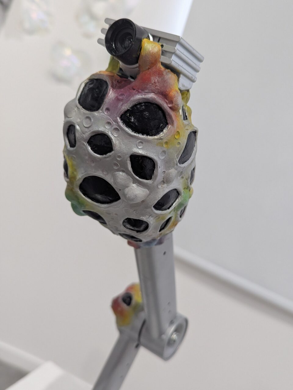 Organic sculptural form with black cavities and coloured surface details attached to a metal support.