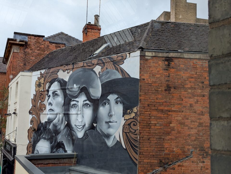Street view of a large monochrome mural of faces framed by brown floral decoration on a brick building.